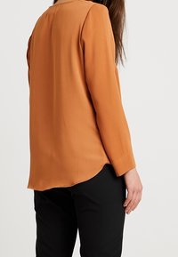 Blusa a maniche lunghe in arancione bruciato, con scollatura rotonda e orlo curvo, abbinata a pantaloni neri. Tessuto dalla texture liscia.