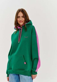 Zelená mikina s kapucňou a ružovými bočnými panelmi, má predné vrecko, ružové šnúrky a malý vyšitý detail v tvare rias.
