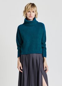 Tiefgrüner Strickpullover mit hohem, gerolltem Rollkragen, cropped Design und geripptem Muster, kombiniert mit einem grauen Faltenrock mit seitlichem Schlitz.