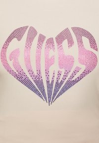 Camisa beige con un gran logo "GUESS" en piedras brillantes de gradiente, que va del rosa al púrpura, con un diseño en forma de corazón.