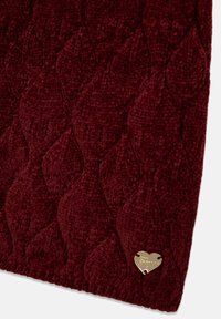 Tissu texturé bordeaux avec un motif de feuilles, doté d'une petite étiquette métallique en forme de cœur doré attachée. Surface douce et moelleuse.