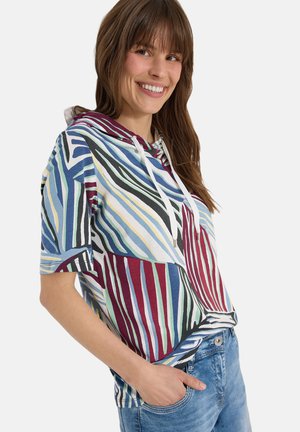 Femme souriante, portant un sweat à capuche rayé multicolore à manches courtes avec des cordons blancs et un jean bleu clair, main dans la poche, sur fond blanc.