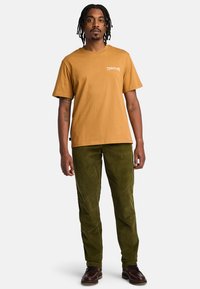 T-shirt a maniche corte color senape con logo bianco "Timberland", abbinato a pantaloni in corduroy verde e scarpe marroni scure. Design semplice e casual.