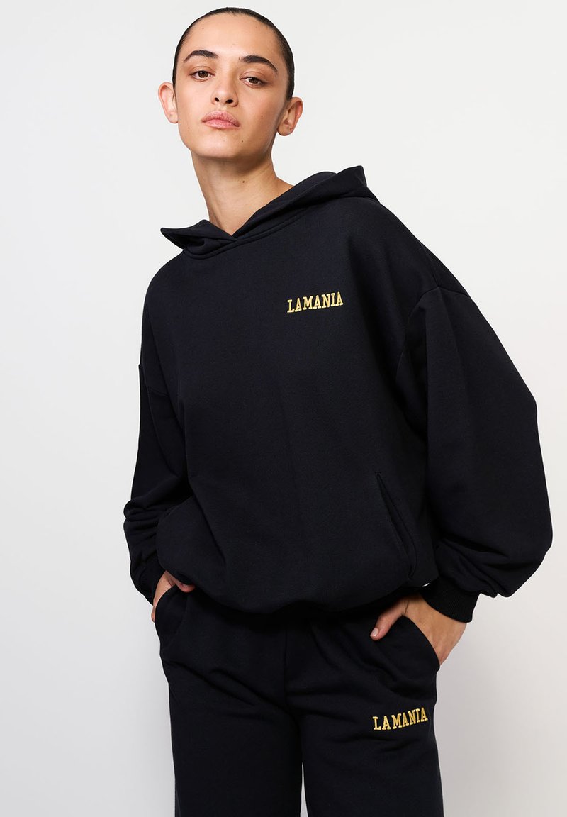Čierna mikina s kapucňou v oversize strihu, s žltou výšivkou "LAMANIA" na hrudníku. Mäkká textúra a bočné vrecká sú súčasťou.