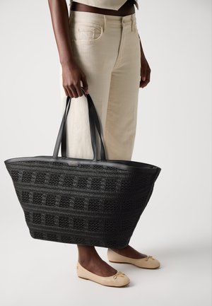 Black woven tote bag with leather trim and handles, featuring a grid pattern. Paired with beige pants and matching ballet flats.