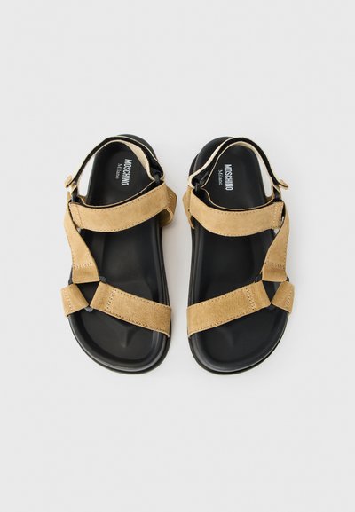 Dvojice černých sandálů na ploché podrážce s nastavitelnými hnědými semišovými popruhy a značkou MOSCHINO Milano na stélkách.