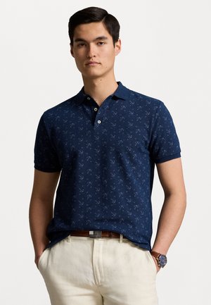 Polo de culoare bleumarin, cu un model subtil de ancoră, mâneci scurte și guler, asortat cu pantaloni de culoare deschisă.
