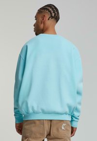 Felpa leggera azzurra a girocollo con polsini e orlo a coste, realizzata in tessuto morbido. Presenta un logo sul lato destro di pantaloni cargo beige.