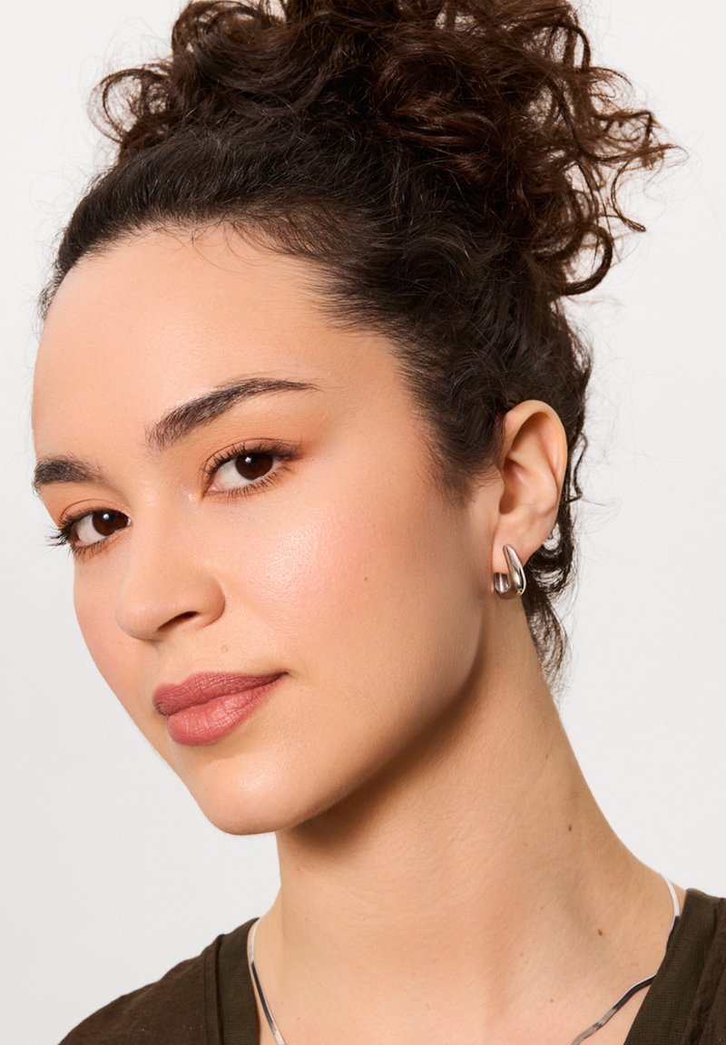 Srebrne kolczyki w kształcie kółek z gładkim, polerowanym wykończeniem; noszone na uchu modelki. Naturalny makijaż i teksturowane, kręcone włosy dopełniają całość.