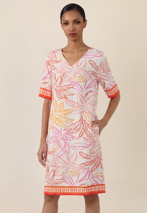 Frau trägt ein knielanges weißes Kleid mit Blättermustern in Pink, Orange und Gelb, V-Ausschnitt und kurzen Ärmeln mit orangefarbenem Saum.