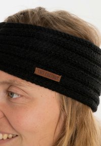 Fascia nera in maglia con design testurizzato, con etichetta in pelle marrone contrassegnata "KNIT FACTORY." Si avvolge attorno alla testa con una fascia spessa.