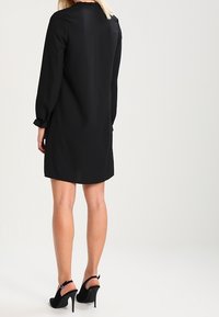 Schwarzes Langarmkleid mit geradem Schnitt, glattem Stoff und minimalistischen Design. Kombiniert mit schwarzen High Heels und Knöchelriemen.