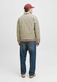 Mand iført en rød cap, beige jakke, ternet skjorte, løse blå jeans og lysebrune sko står vendt væk mod en ensfarvet baggrund.