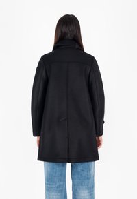 Cappotto nero con colletto strutturato, maniche lunghe e forma a trapezio. Tessuto liscio con cuciture visibili e dettaglio con bottoni sul retro.