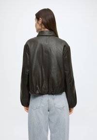 Casaco de couro castanho escuro com design curto, apresenta gola alta, bainha elástica e mangas largas. Usado sobre jeans azul claro.