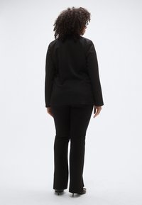 Fekete, testre szabott blézer és nadrág hátulnézetből. Az anyag simának tűnik, szabott fazonú, minták nélkül. Magassarkú cipőt visel.