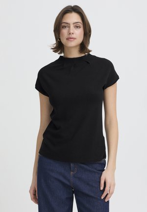 Femeie îmbrăcată într-un tricou negru cu mâneci scurte și blugi albaștri, stând în picioare pe un fundal simplu gri deschis, cu fața spre înainte.