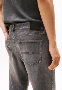 Person wearing faded black denim jeans with logo patch on back pocket and a dark gray t-shirt tucked in.