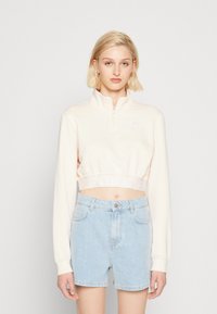 Svetloružový skrátený pulóver s štvrť-zipsom, s bielym logom na hrudníku a elastickým pásom na spodnom okraji. V kombinácii so svetlomodrými džínovými šortkami.