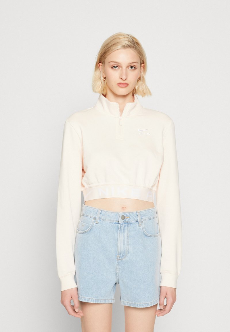 Svetloružový skrátený pulóver s štvrť-zipsom, s bielym logom na hrudníku a elastickým pásom na spodnom okraji. V kombinácii so svetlomodrými džínovými šortkami.