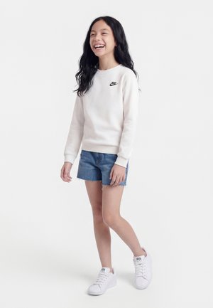 Jeune fille souriante, portant un sweat-shirt blanc Nike, un short en jean et des baskets blanches, debout devant un fond uni.