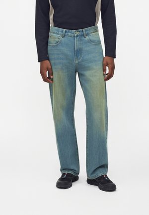 Personne portant un jean bleu délavé droit, des baskets noires et un t-shirt noir à manches longues avec des panneaux latéraux beige, debout sur fond blanc.