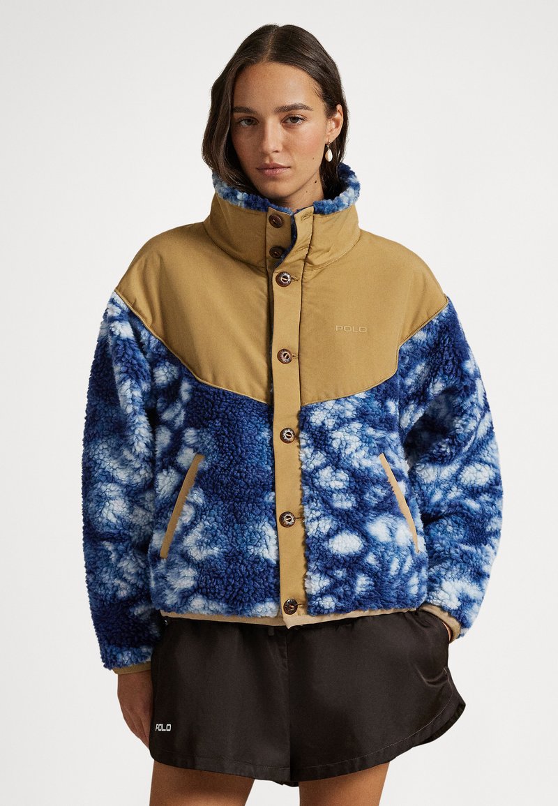 Ζακέτα φλις με πάνω μέρος σε ταμπά  απόχρωση και κάτω μέρος με μπλε εφέ tie-dye, με ψηλό γιακά, κουμπιά μπροστά και πλαϊνές τσέπες. Μαύρο σορτς με λογότυπο από κάτω.