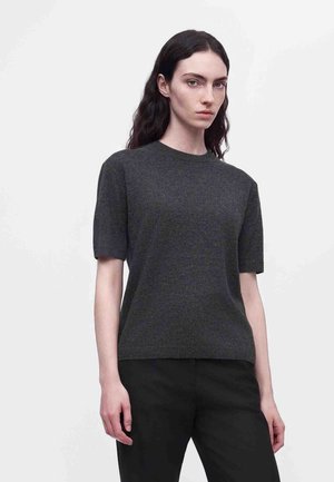 Un mannequin aux longs cheveux foncés porte un haut en maille gris anthracite à manches courtes et un pantalon noir sur un fond clair uni.