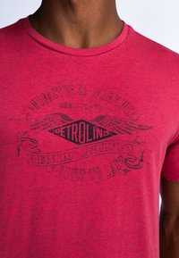 Červené tričko s krátkým rukávem a kulatým výstřihem, se černým grafickým textem a vzory ve středu. Vyrobeno z texturované, melírované látky.