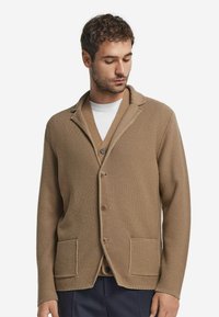 Beige stickad kofta med sjalkrage, knappstängning och två framfickor. Har ett strukturerat mönster och synliga knappar.