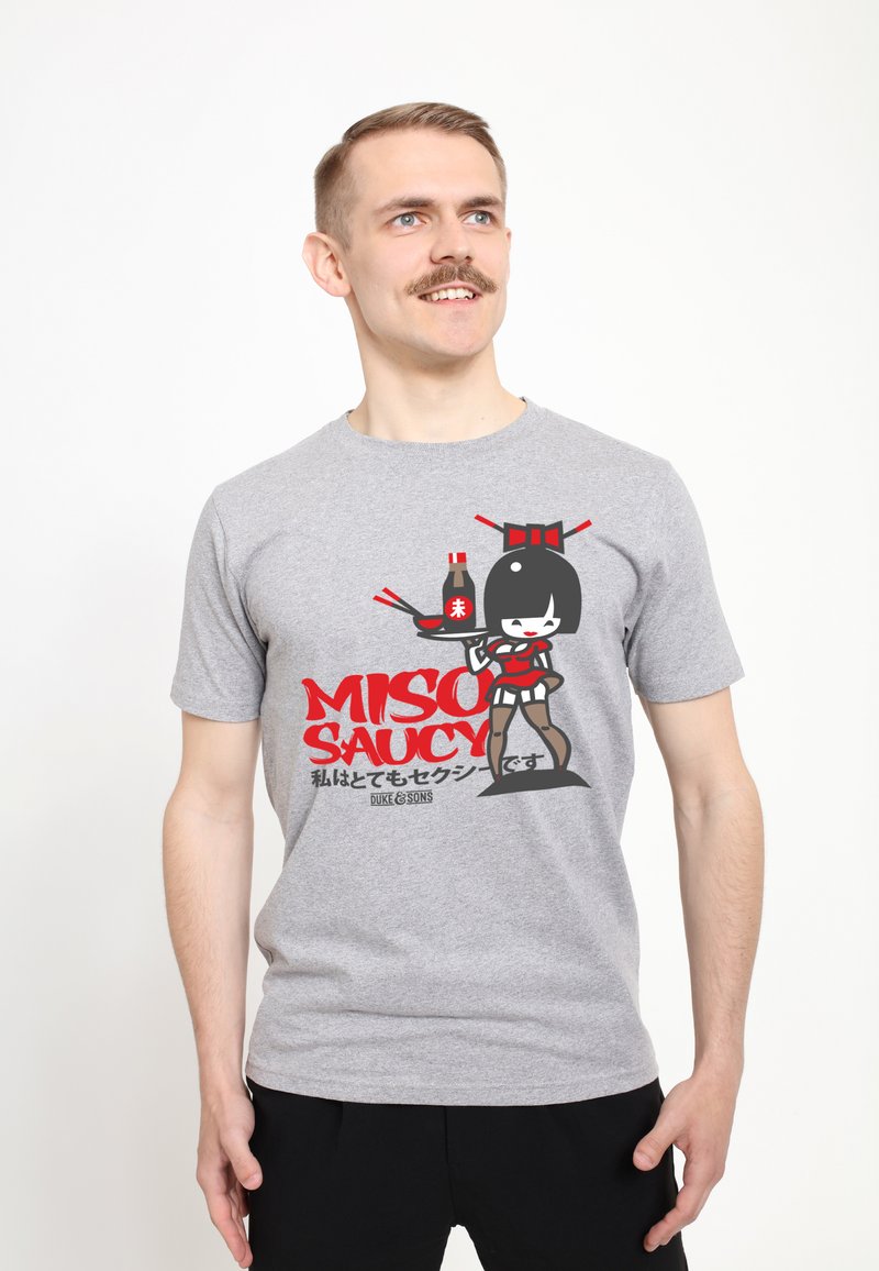 Camiseta de algodón gris con un gráfico de dibujos animados de un personaje sosteniendo salsa de soja, con el texto rojo "MISO SAUCY" y acentos en negro.