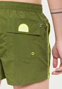 Shorts de bain verts en tissu léger et résistant à l'eau. Dotés d'une poche arrière avec un logo jaune et des coutures jaunes contrastantes.