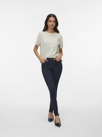 Frau steht mit den Händen in den Taschen, trägt ein weißes und hellblau gestreiftes kurzärmeliges Hemd, dunkelblaue Skinny-Jeans und schwarze High Heels.