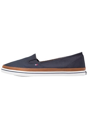 Zapato de lona azul marino tipo slip-on con ribete marrón, suela de goma blanca y un pequeño logo rectangular rojo, blanco y azul en el lateral.