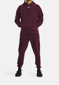 Bordeauxrotes Hoodie- und Jogginganzugset aus weichem Stoff. Verfügt über eine Fronttasche, gerippte Bündchen und ein gesticktes Logo auf der Brust. Schwarze Sneaker.