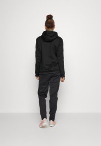 Haut à capuche noir au design ajusté, associé à un pantalon de jogging noir. Il présente une capuche réglable, des poignets côtelés et des chaussures de sport grises.