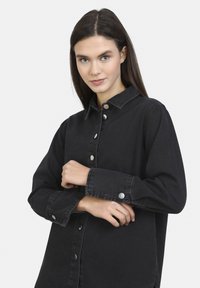La camicia in denim nera presenta una chiusura con bottoni sul davanti e un colletto, con dettagli in metallo argentato e maniche lunghe, che mette in risalto un taglio rilassato.