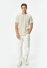 Beige kortærmet T-shirt, der er parret med hvide jeans. Stoffet ser glat ud, og pasformen er afslappet. Fodtøjet er lysfarvede sneakers.