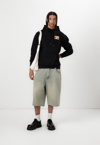 Svart sweatshirt med framficka och logotyp, ljus tvättade denimshorts, vita strumpor, svarta klumpiga skor och en krämfärgad tote-väska.