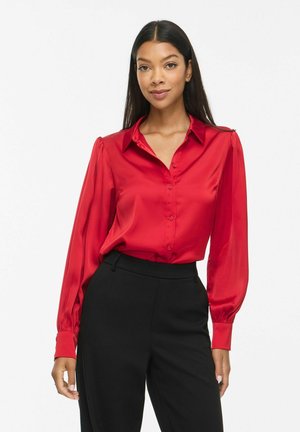 Femme aux longs cheveux foncés vêtue d'un chemisier rouge boutonné avec des manches bouffantes et d’un pantalon noir taille haute, debout devant un fond blanc.