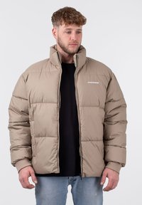 Beige Steppjacke mit hohem Kragen, gepolsterten Abschnitten und einem Frontreißverschluss. Verfügt über ein Logo auf der Brust und gerippte Bündchen. Trägt man über einem schwarzen Hemd.