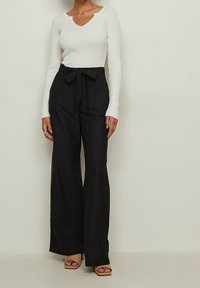 Witte geribbelde lange mouw top met een V-hals en zwarte high-waisted broeken met een strikceintuur. Model draagt naakte blokhak sandalen.