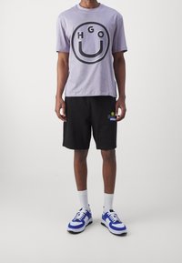 Lilla bomulds t-shirt med et stort sort cirkulært grafisk motiv med "HGO". Sammen med sorte shorts og blå og hvide sneakers.