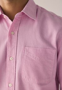 Svijetlo ružičasta košulja na dugih rukava od teksturirane tkanine, s klasičnim ovratnikom i jednim džepom na lijevoj strani grudi s suptilnim detaljima šivanja.
