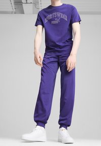 Purple cotton t-shirt with "SPORTSWEAR PUMA" lettering, paired with matching purple sweatpants and white sneakers, featuring elastic cuffs.