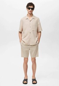 Lichtbeige shirt met korte mouwen, enkele borstzak, gecombineerd met lichtgestreepte shorts. Zwarte zonnebril en zwarte sandalen maken de look compleet.