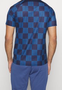 Blå mönstrad träningsskjorta med rutigt mönster, med upprepande text på en mörk marin bakgrund. Kortsleeve och vanlig passform.