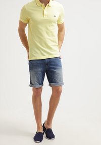 Polo jaune, short en jean avec revers, et chaussures bleu marine sans lacets. Logo emblématique sur la poitrine gauche. Tenue d'été décontractée.