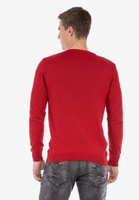 Roter Strickpullover mit langen Ärmeln und rundem Ausschnitt, versehen mit einem strukturierten Muster. Getragen mit dunkelgrauen Jeans, von hinten betrachtet.