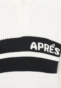 Πλεκτός πουλόβερ σε λευκό και μαύρο, με ψηλό γιακά, κεντρικό φερμουάρ και κείμενο "APRÈS" με οριζόντιες ρίγες. Υφασμάτινη υφή.